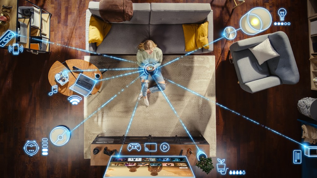 A person is sitting in a living room, with a mobile device in their hands. Blue lines connect the mobile device to items in the person's home, indicating wireless connectivity between the mobile device and home items. The home items include a light fixture, a thermostat, a plant, a TV, a robot vacuum, a computer, headphones, and a stereo system.